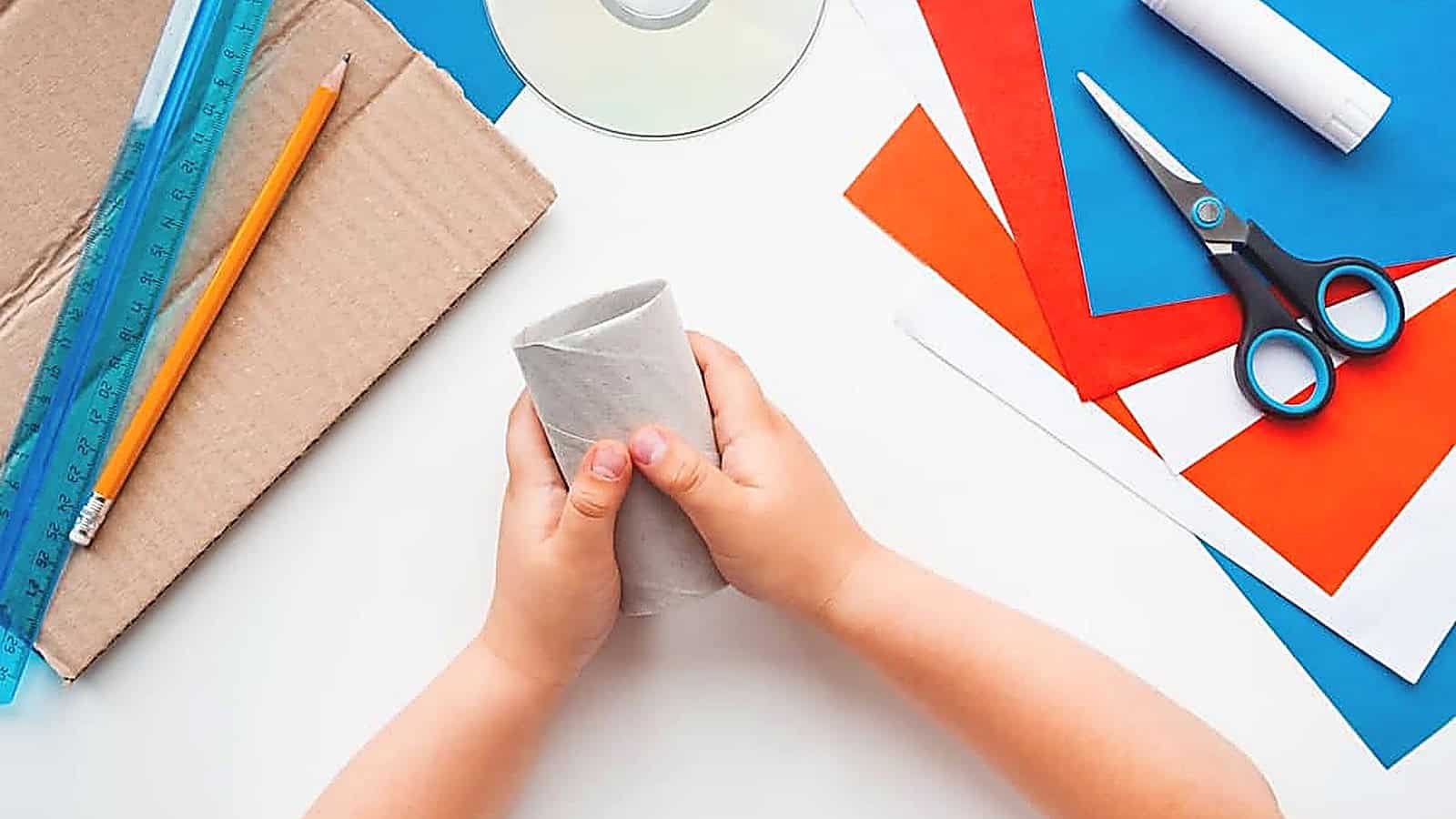 Child’s hands holding a cardboard tube on a table with colored paper, scissors, glue stick, ruler, pencil, a CD, and a piece of cardboard, ready to create fun and eco-friendly toys in this imaginative craft project.