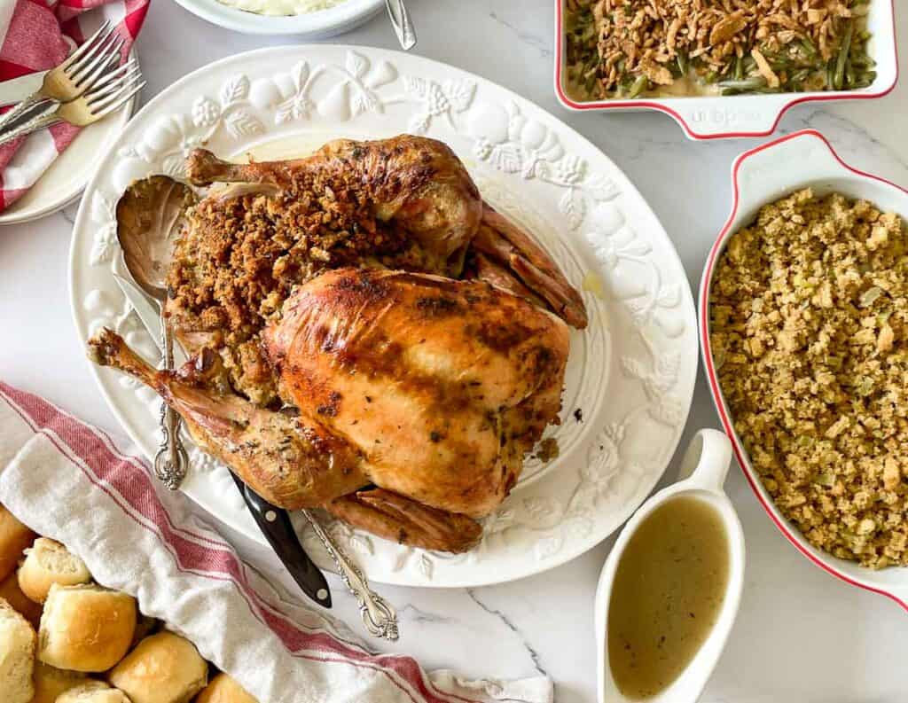 Roast turkey with stuffing on a white platter, surrounded by bowls of green bean casserole, stuffing, rolls, and a gravy boat on a white table.