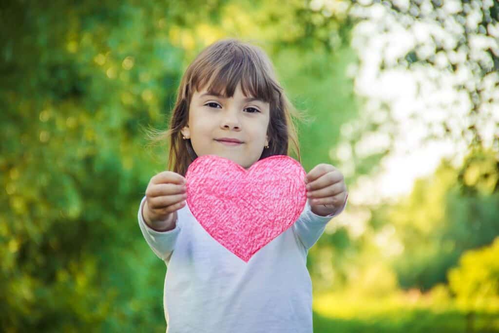 A little girl is holding up a pink heart.