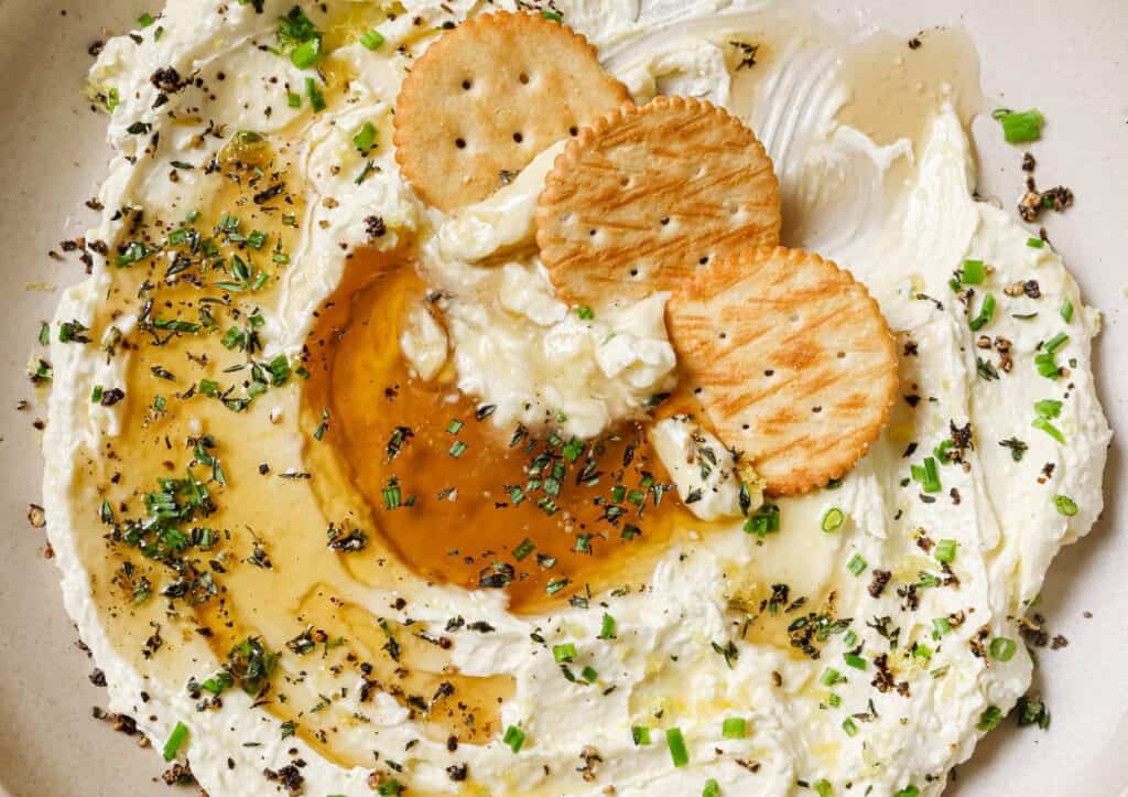 A plate of whipped feta with honey dip with crackers and herbs.