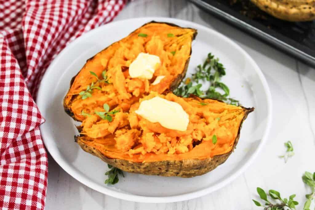 A baked sweet potato split in half on a white plate, topped with butter and herbs, sits next to a red checkered cloth&mdash;an easy air fryer recipe for a quick, delicious side.