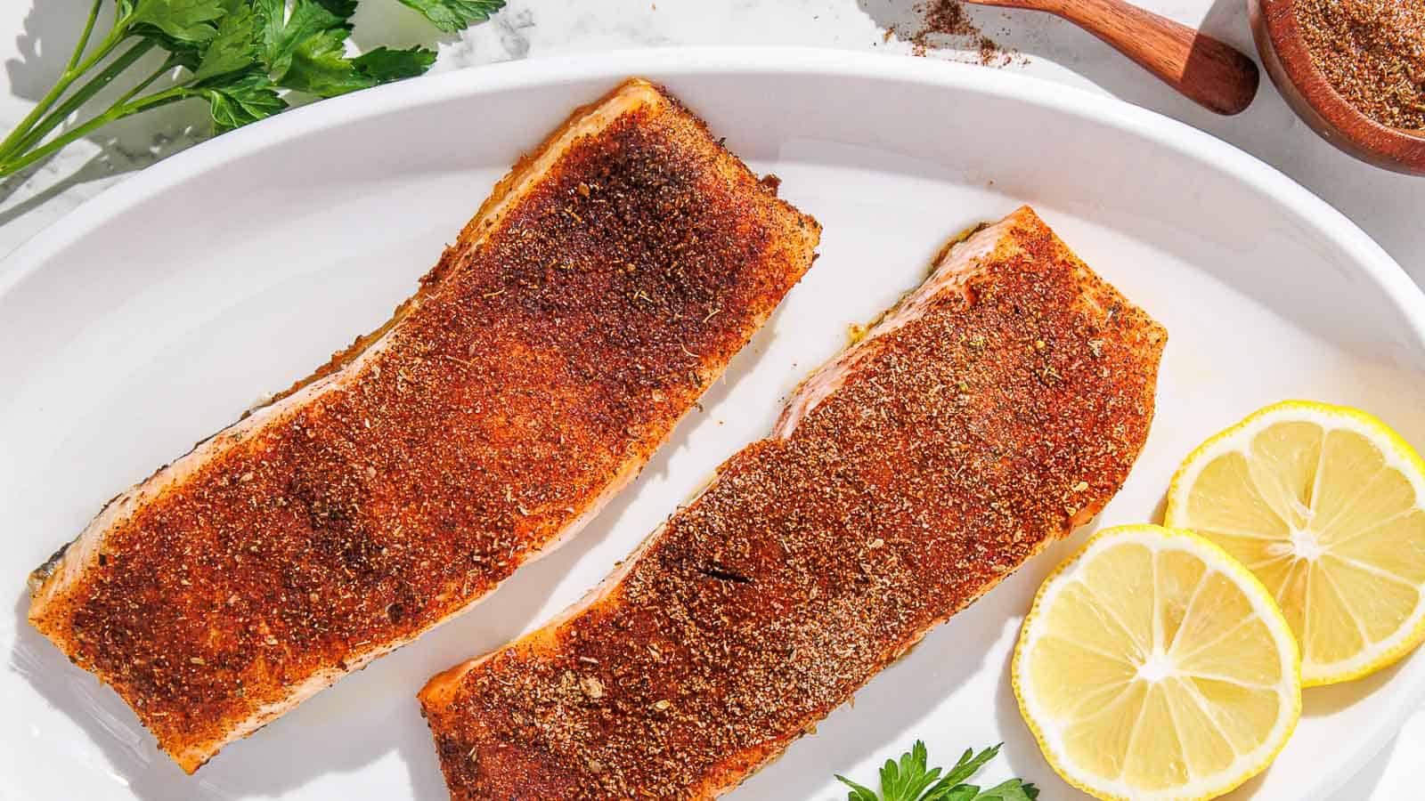 Two blackened salmon fillets on a plate with lemon and parsley.