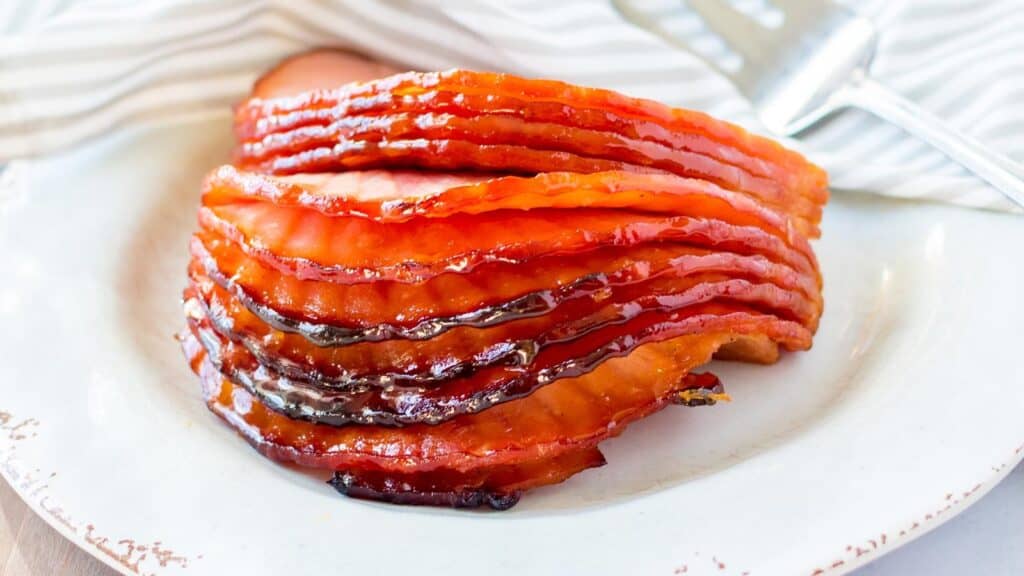 Sliced maple mustard glazed ham on a white plate with a towel in the background. 