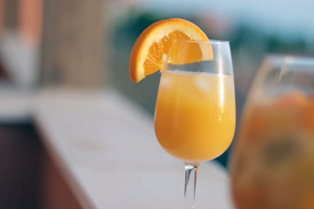 A glass of orange juice mimosa with a slice of orange sitting on a balcony.