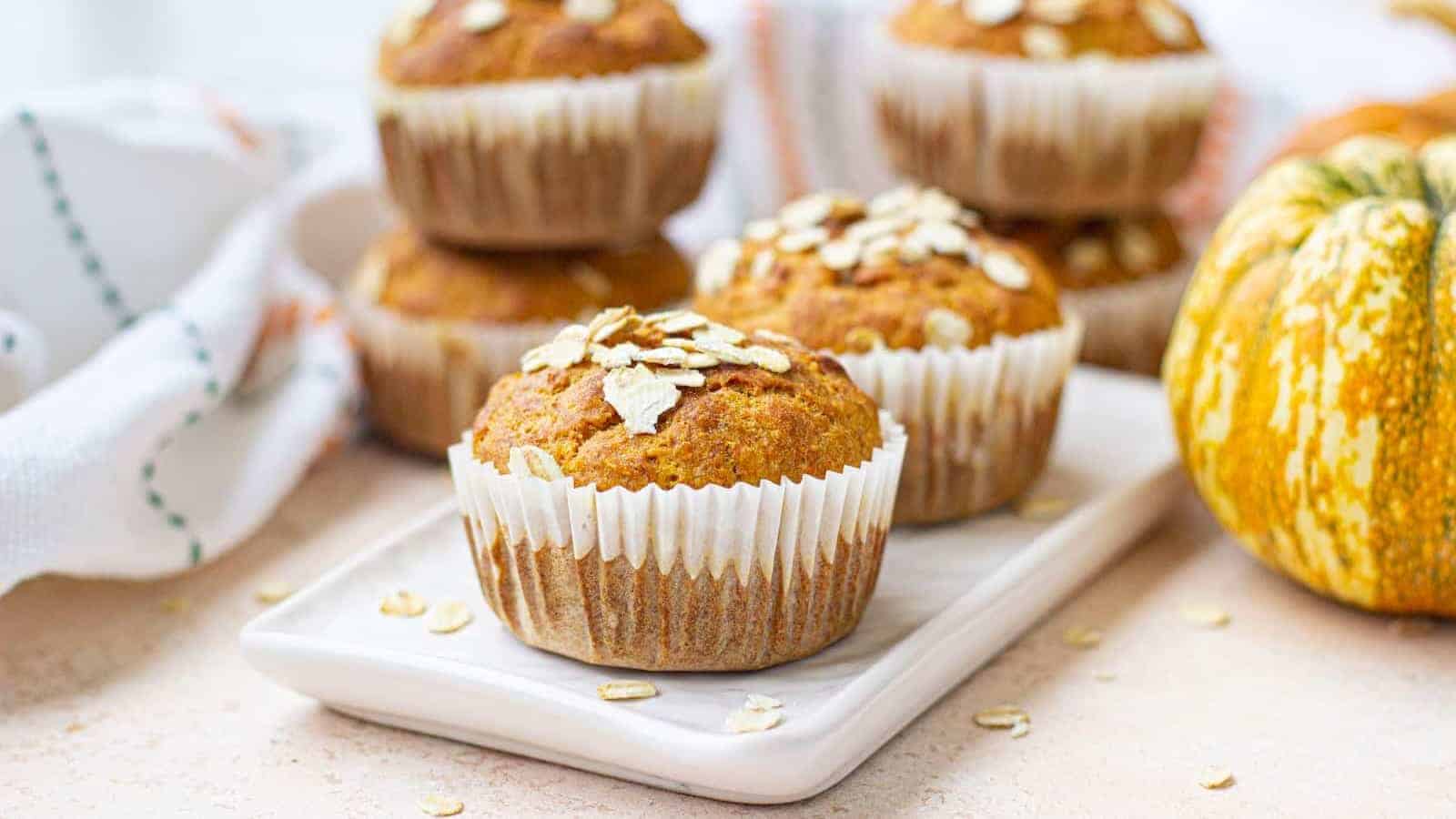Pumpkin banana oat muffins on a white plate with pumpkins in the background.