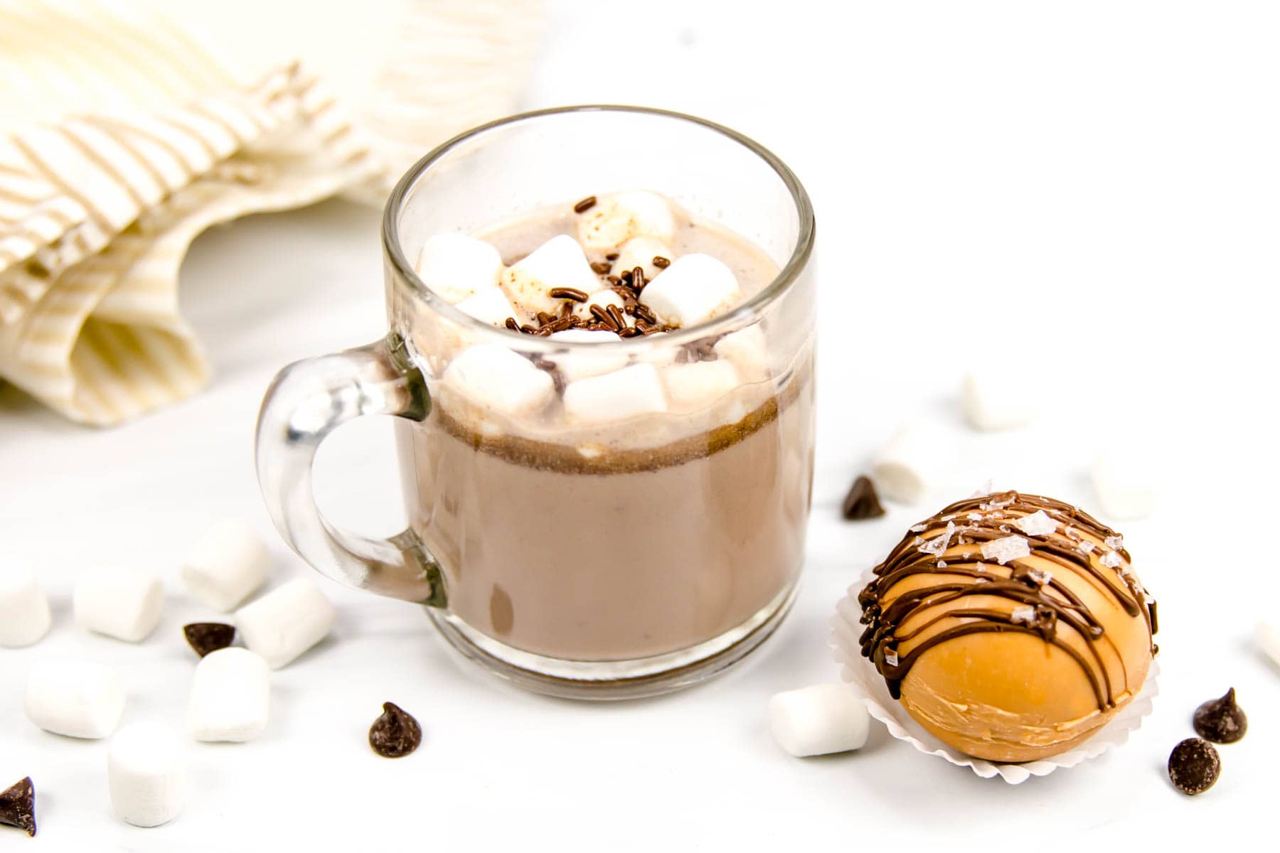 A mug of salted caramel hot chocolate sits next to a salted caramel hot chocolate bomb, marshmallows and chocolate chips.