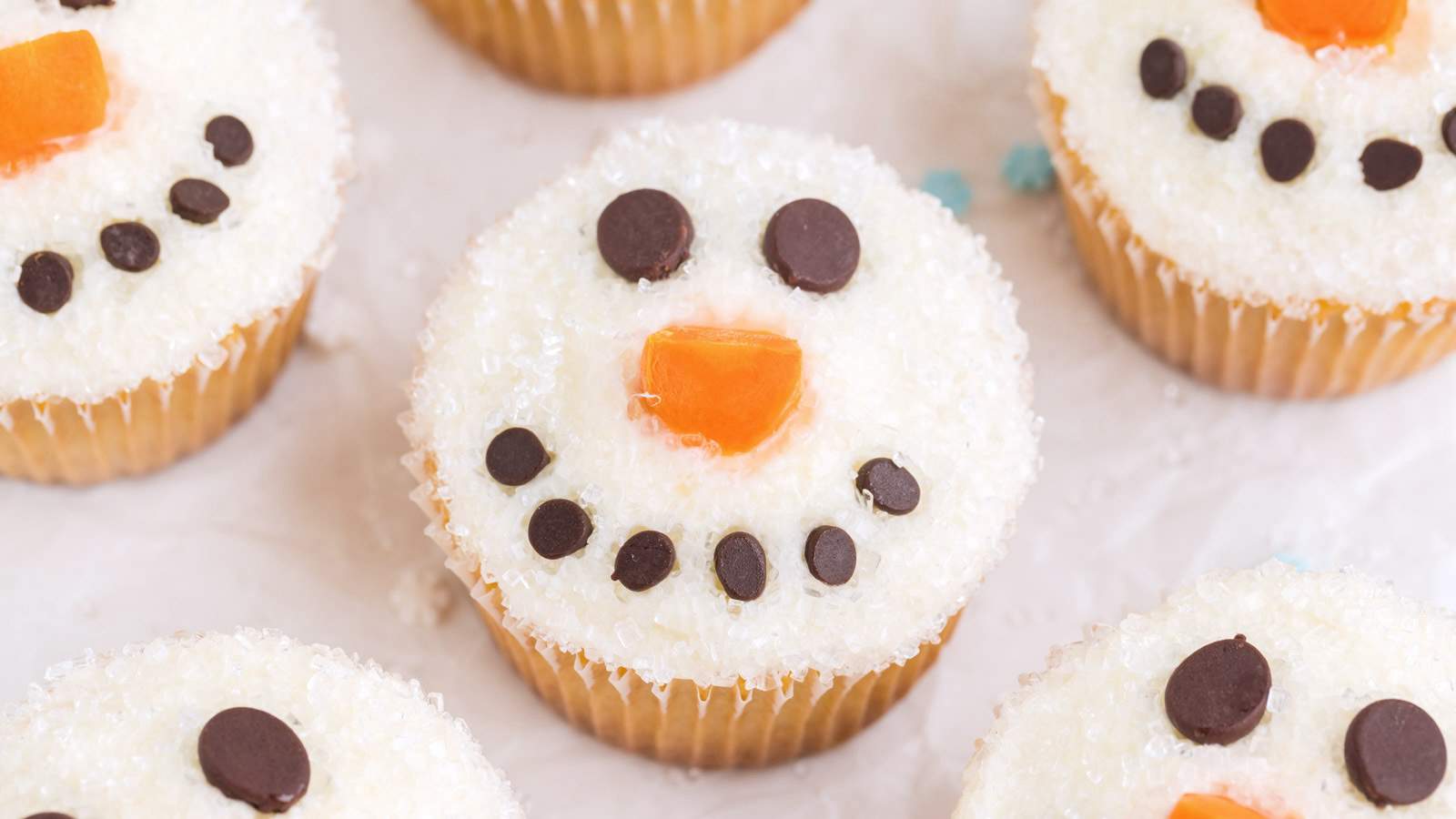Snowman Cupcakes on a white piece of parchment paper with snowflake sprinkles in the background.