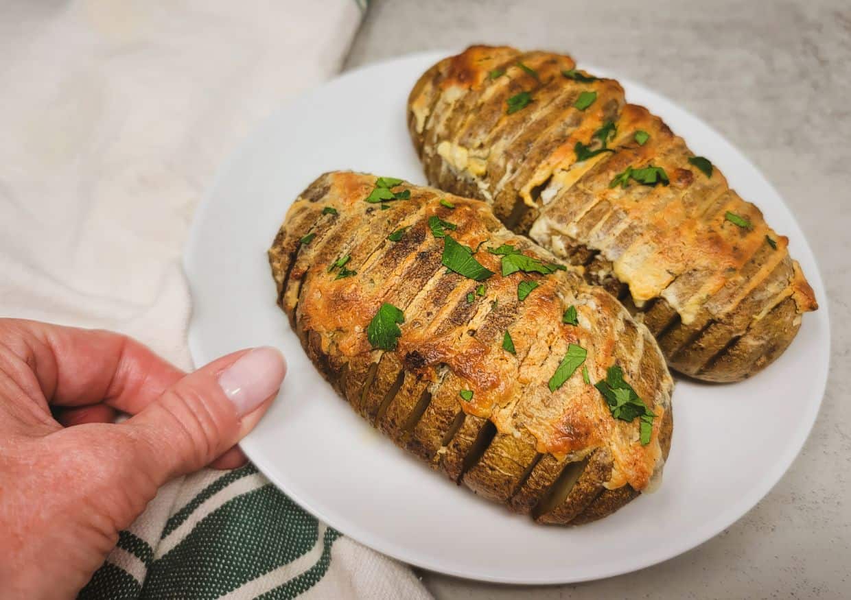 Two hasselback style potatoes topped with baked Boursin on a white plate with a hand holding them.