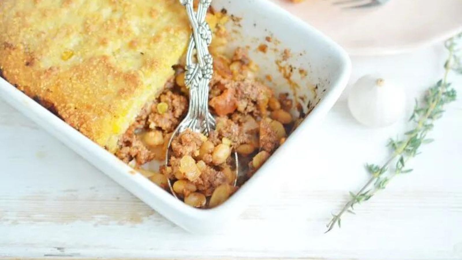 A white casserole dish with cornbread casserole and a silver serving spoon.