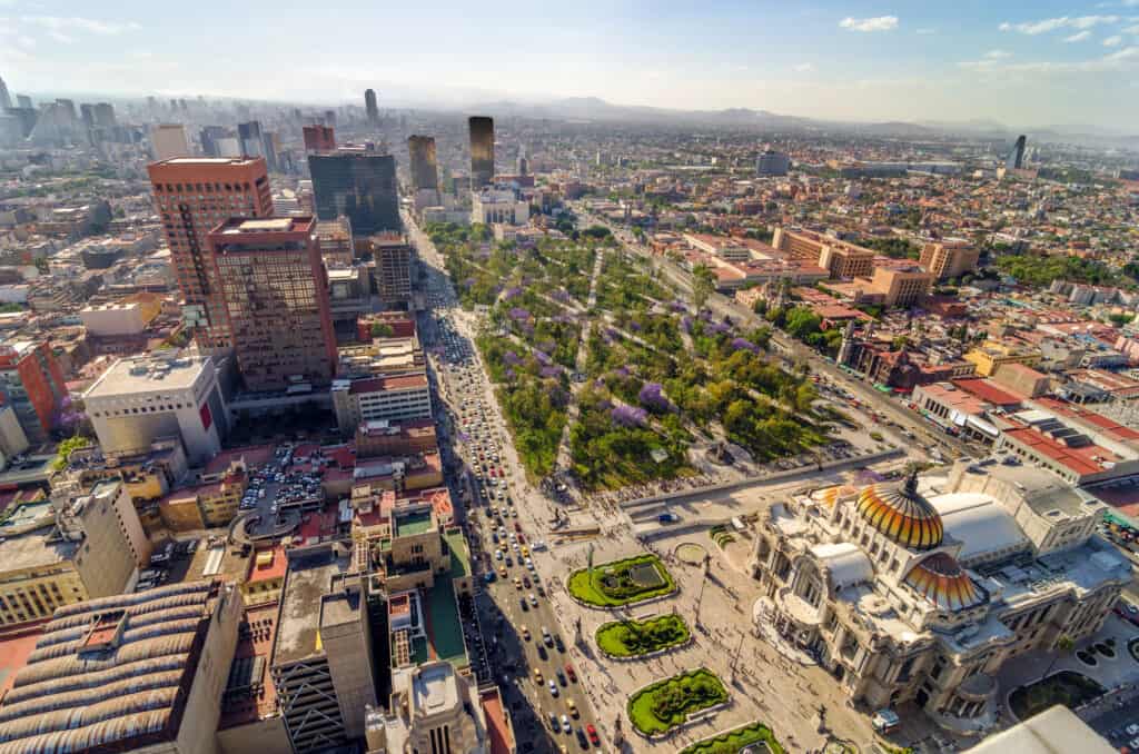 Aerial view of Mexico City Travel highlights a cityscape with a large park lined with purple-blossomed trees, busy streets, and a mix of historic and modern buildings under a clear sky.