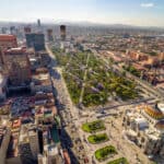 Aerial view of Mexico City Travel highlights a cityscape with a large park lined with purple-blossomed trees, busy streets, and a mix of historic and modern buildings under a clear sky.