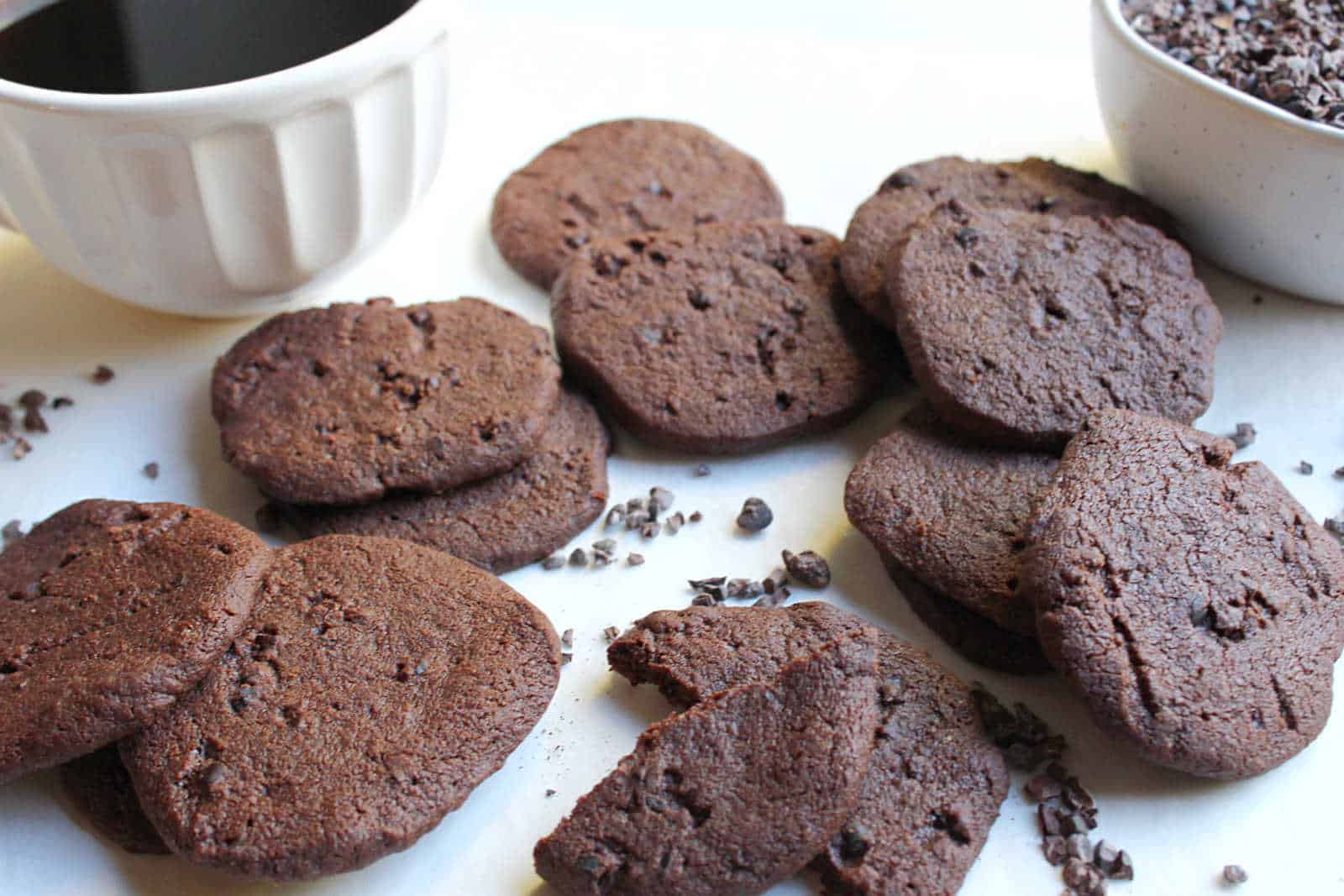 A group of chocolate cookies is arranged on a surface near a white cup of coffee and a bowl of cocoa nibs. One cookie is partially bitten.