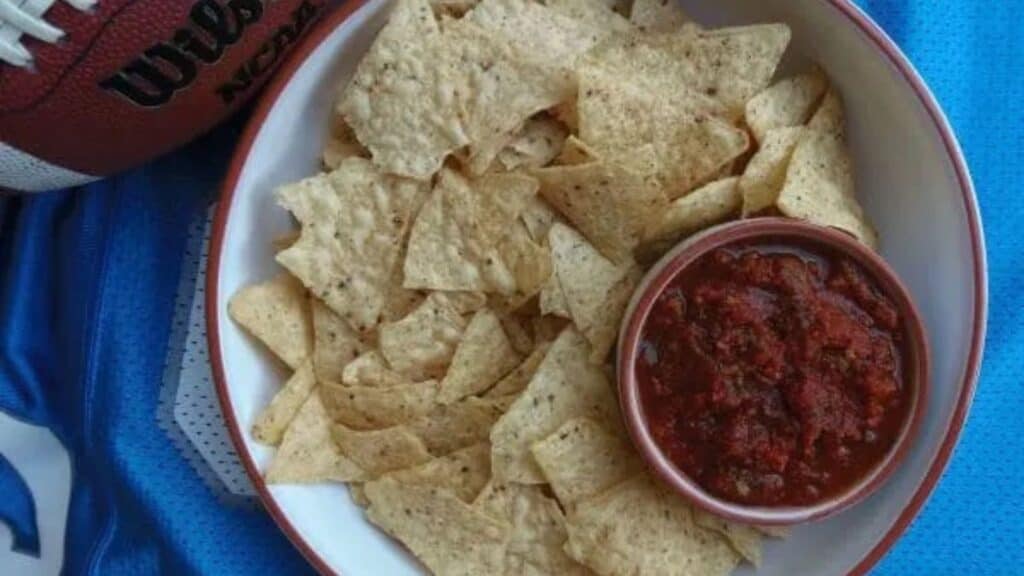 A bowl of nachos and a football on a blue blanket.