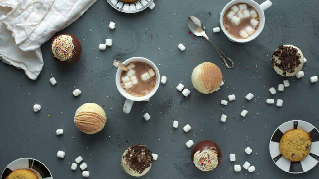 Hot chocolate bombs, marshmallows, mugs of hot chocolate, and cookies on a table.