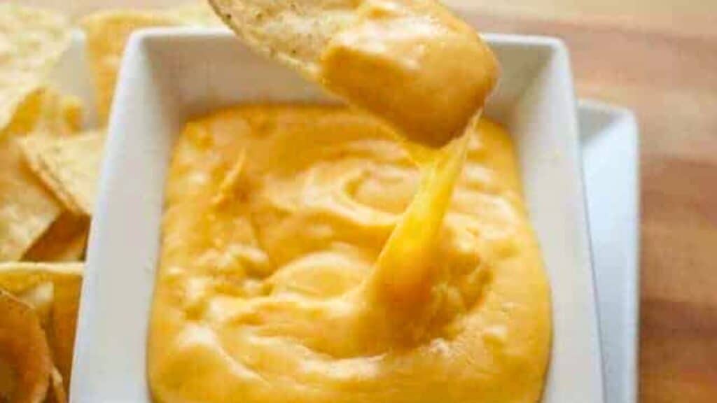 Image shows a tortilla chip pulling nacho cheese out of a white bowl filled with it.