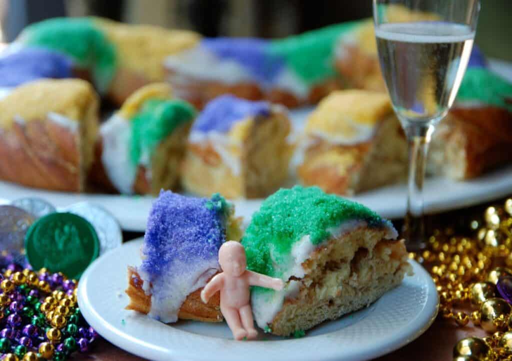 A piece of king cake on a table decorated for Mardi Gras next to a glass of champagne.
