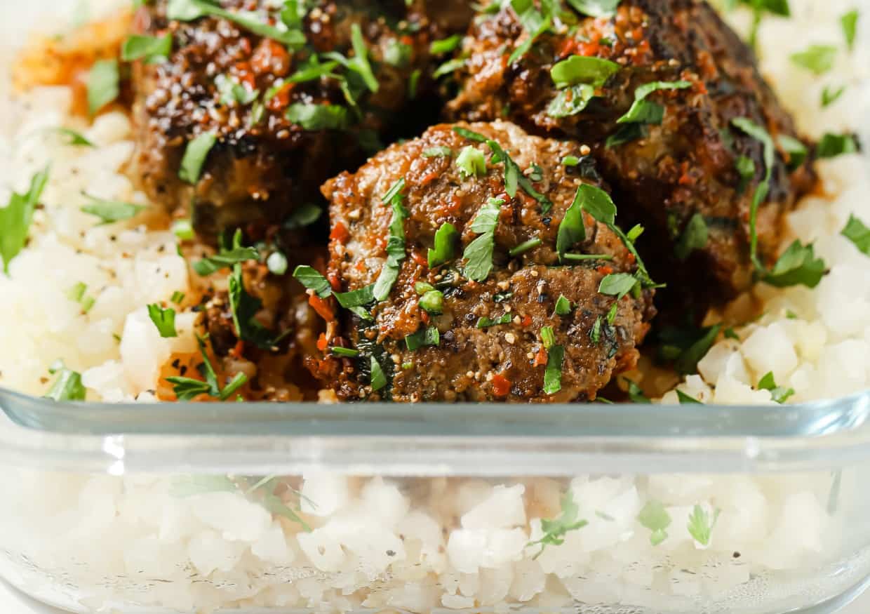 A glass container with garnished meatballs over cauliflower rice.