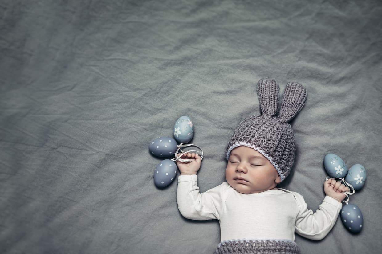 A newborn wearing a knitted bunny hat and holding easter eggs sleeping on grey background.