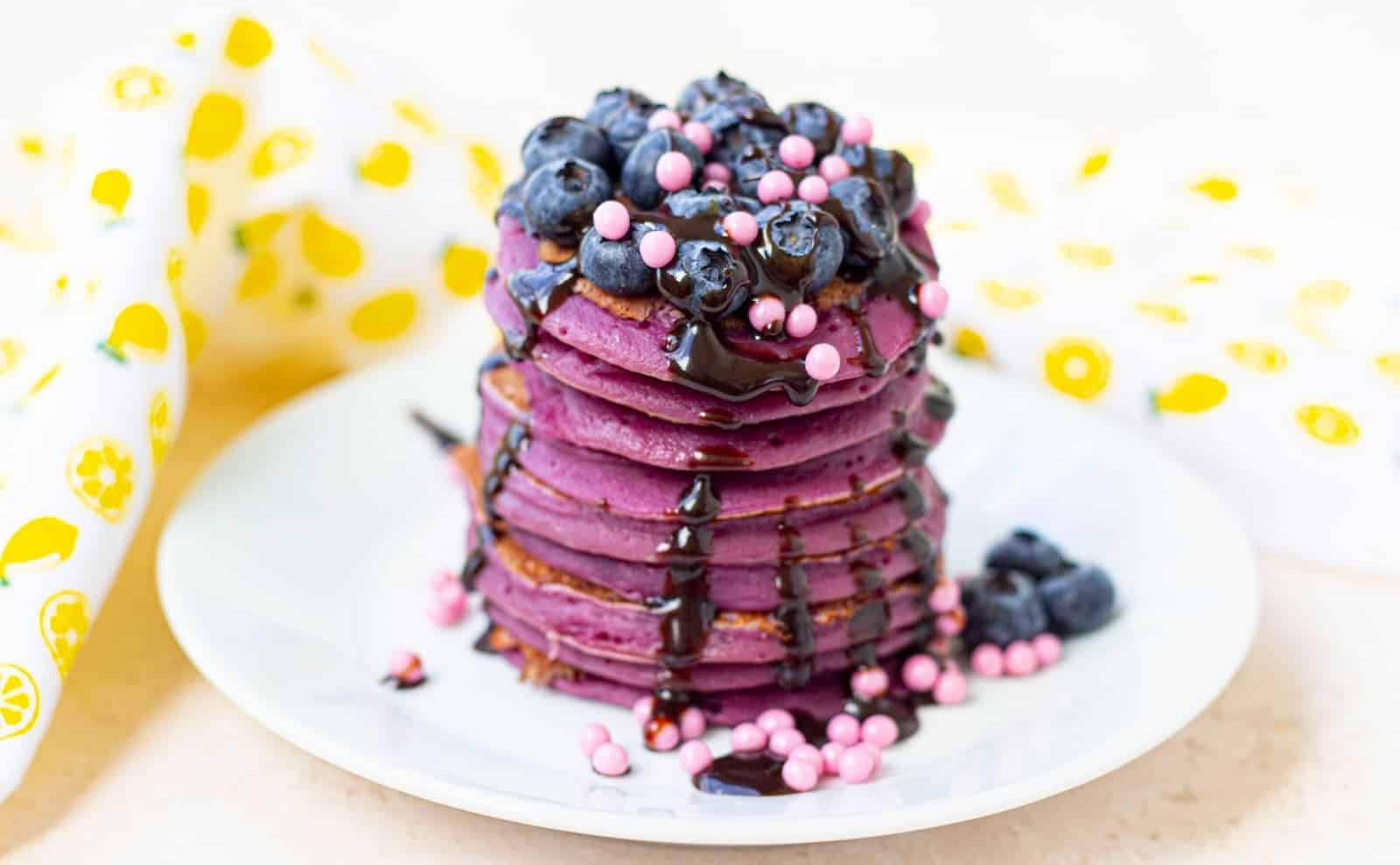 A stack of purple pancakes with blueberries and chocolate sauce on a white plate.