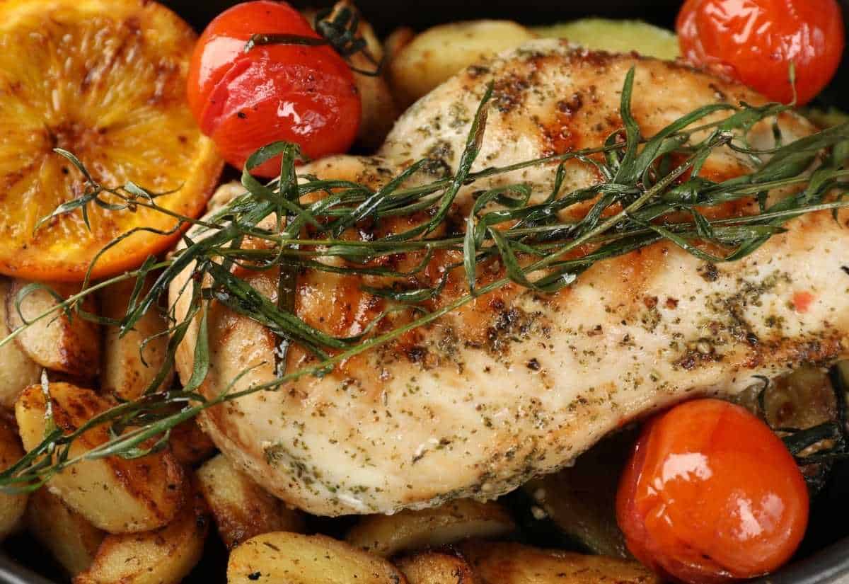 Grilled chicken breast topped with fresh tarragon and herbs, surrounded by roasted potatoes, cherry tomatoes, and a slice of orange.