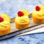 Three cupcakes with lemon icing and raspberries on a tray.