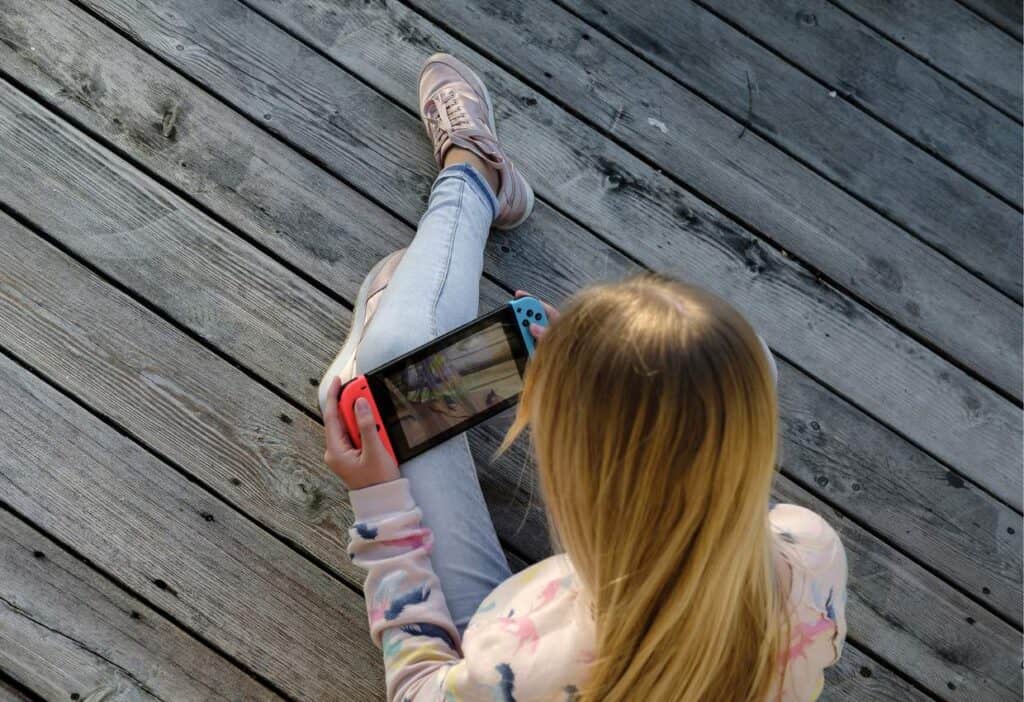 A girl sitting on a wooden deck enjoying a Nintendo Switch tablet while playing games perfect for kids.