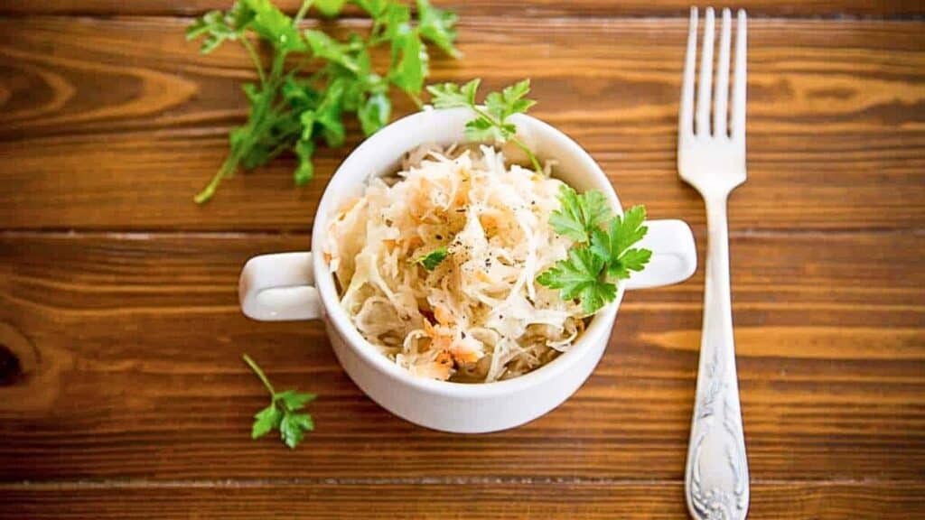 A white bowl of sauerkraut, one of the best natural probiotic foods, is garnished with parsley and sits on a wooden table next to a fork and a few parsley sprigs.