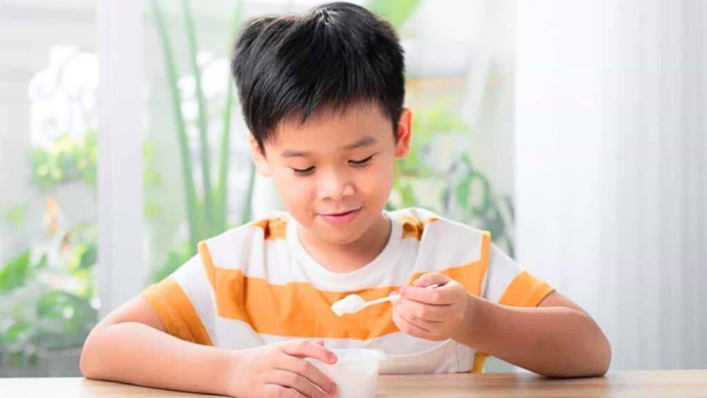 A young boy wearing a yellow and white striped shirt sits at a table, eating yogurt with a spoon&mdash;one of the delicious natural probiotic foods that support digestive health.