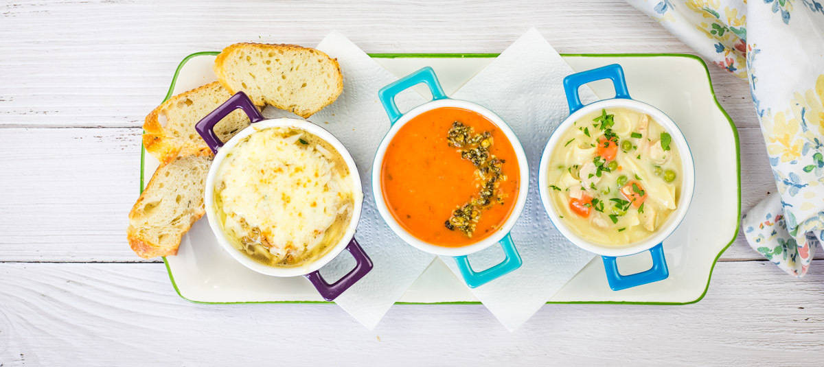 Three bowls of different kinds of soup accompanied by slices of bread served on a white wooden table.