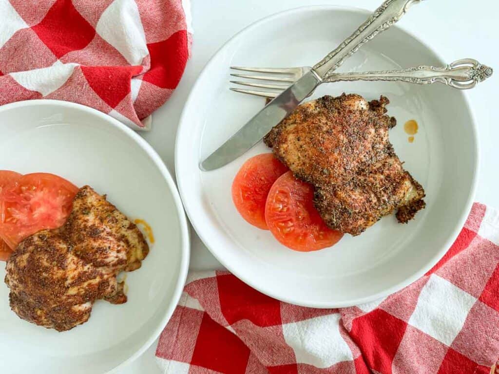 Two plates of chicken with tomatoes and a fork on a checkered tablecloth.