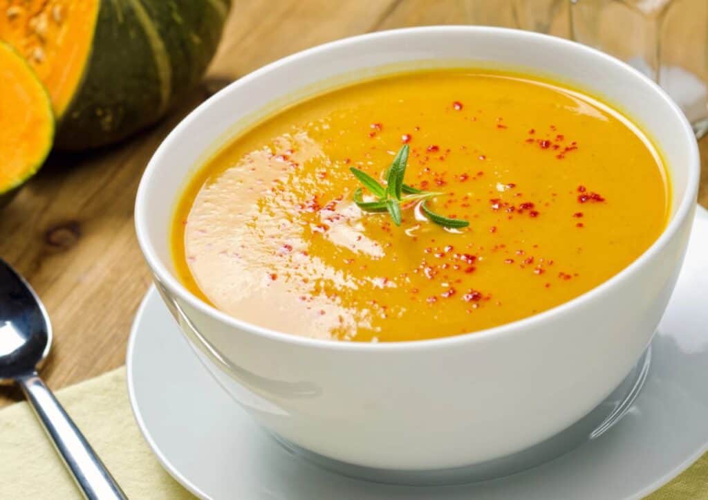 A bowl of pumpkin soup with a spoon on a wooden table, drizzled with maple syrup.