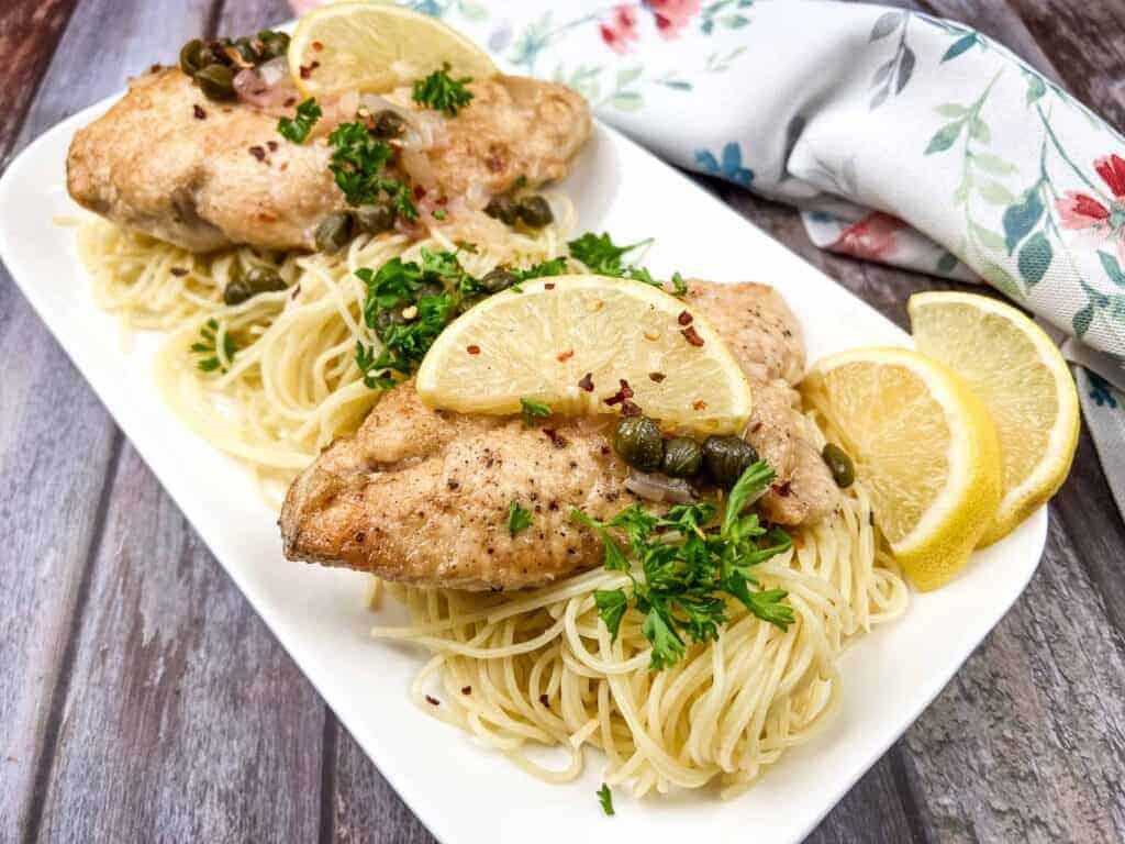 Two chicken piccata on a white plate with parsley and lemon wedges.