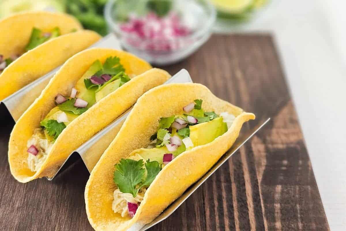 Three chicken tacos topped with cilantro, avocado and red onions on a wooden cutting board.