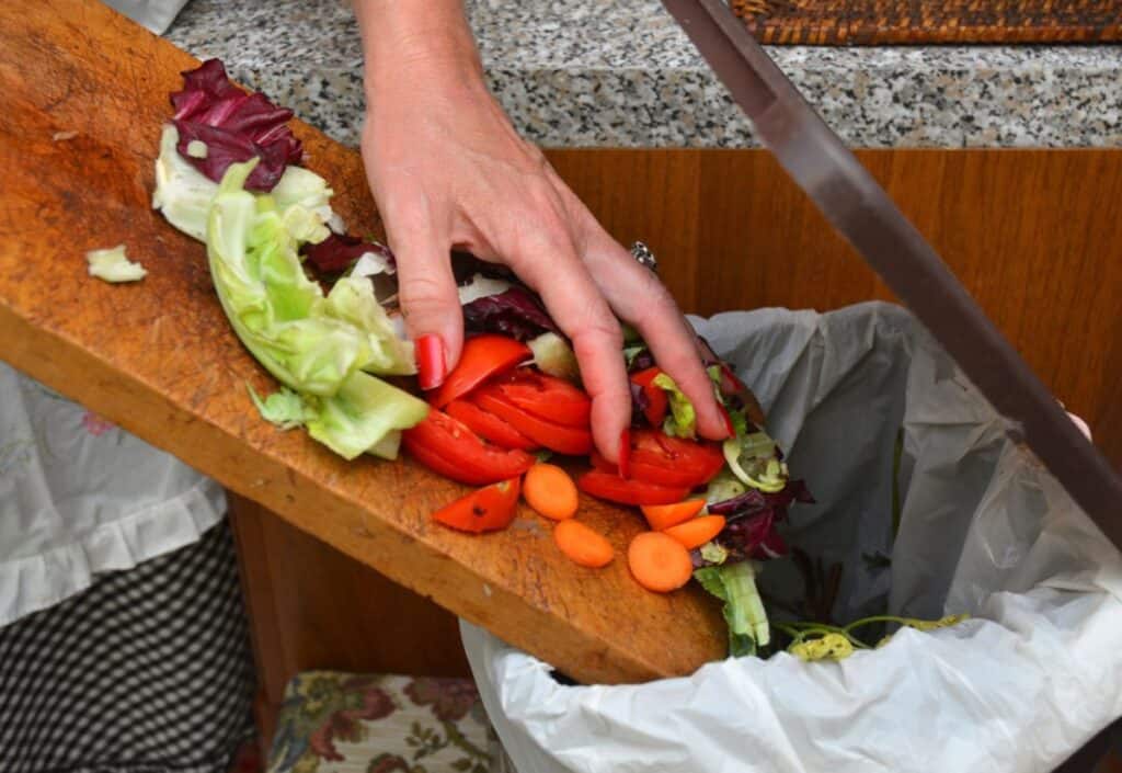 A woman is disposing of food waste by putting vegetables into a trash can.