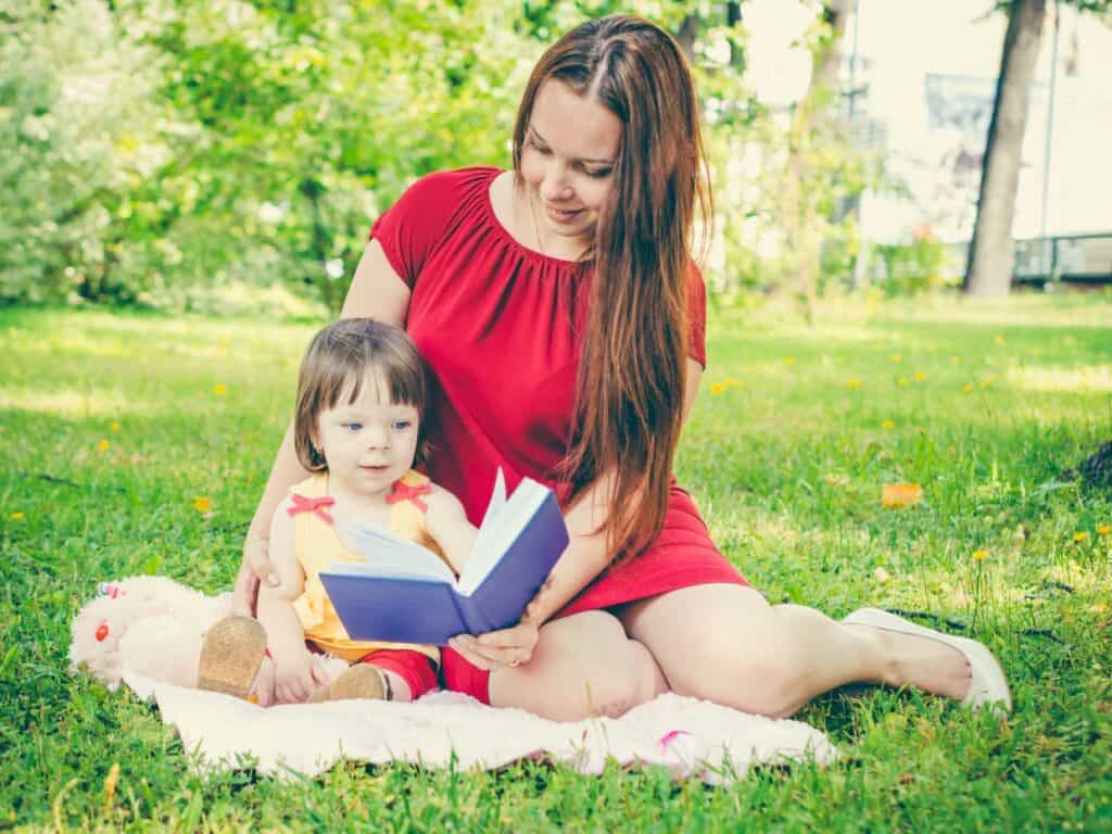 A woman is reading a book to her daughter in a park.