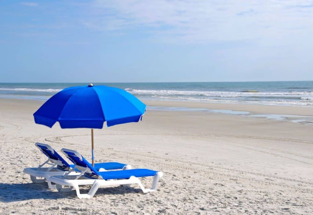 Two blue lounge chairs on the beach with an umbrella.