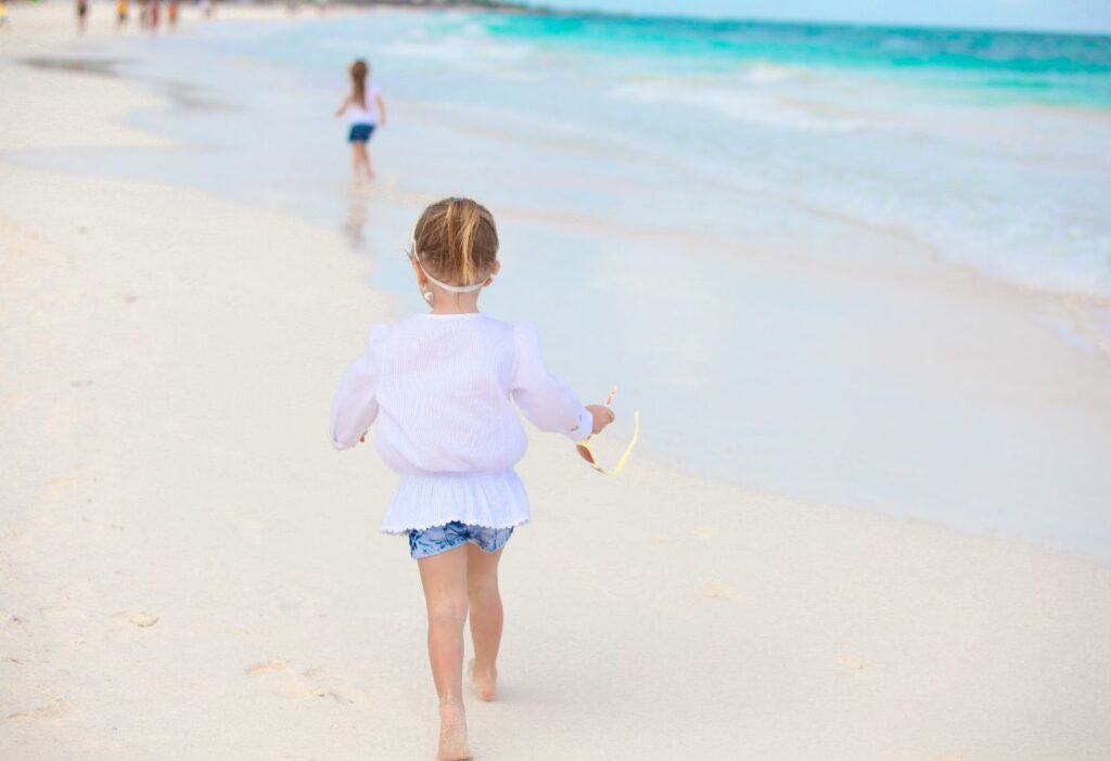 A little girl is walking on the beach with a pair of flip flops.