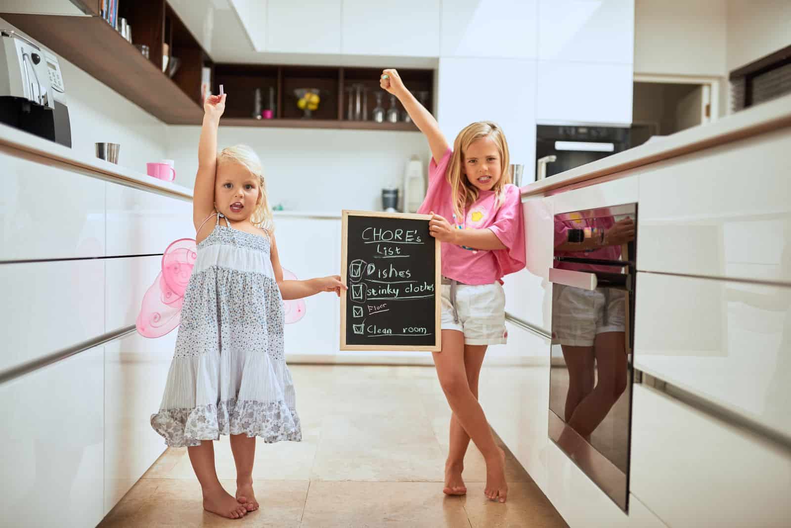 Two little girls standing in a kitchen holding a chalkboard.