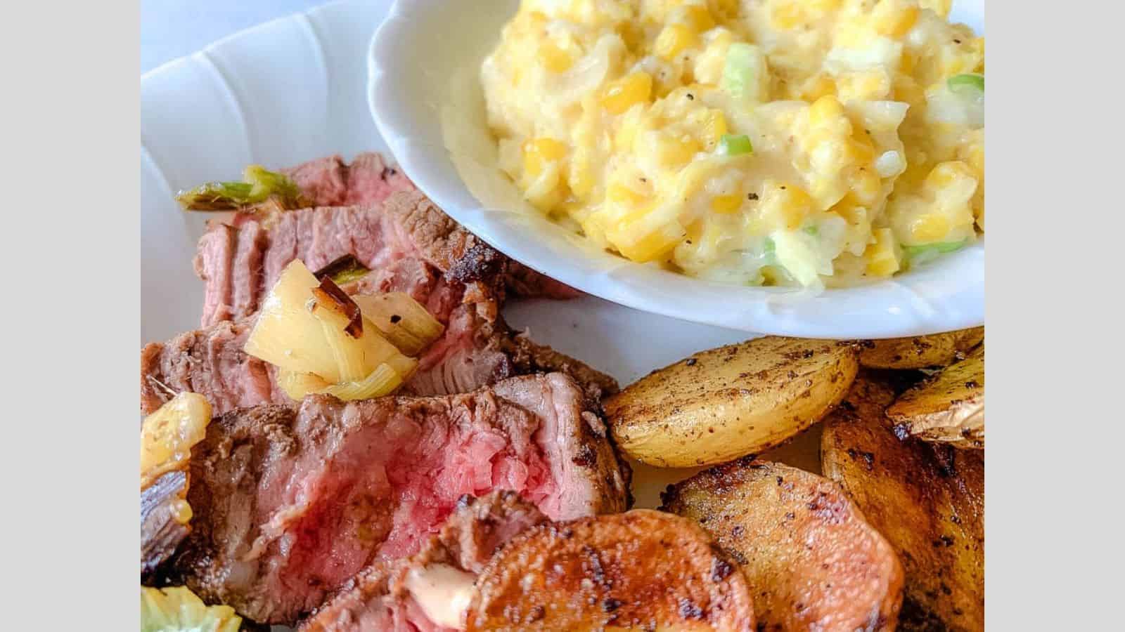 A plate filled with grilled steak, charred onions, potatoes, and creamed corn.