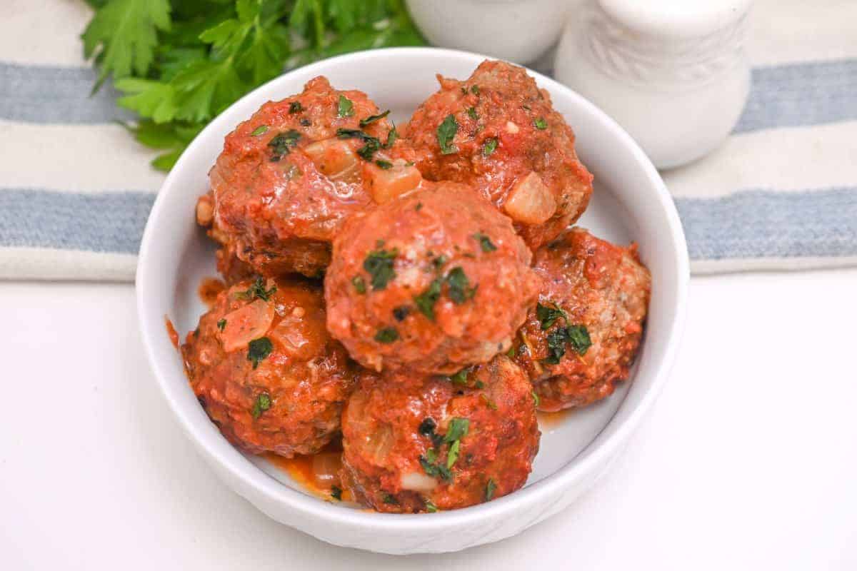 Meatballs in a white bowl with sauce and parsley.