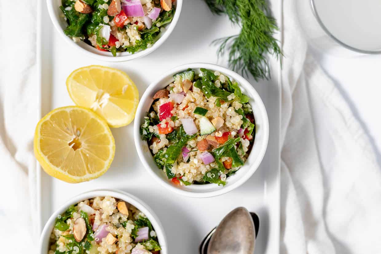 A bowl of kale quinoa salad with fresh sliced lemons and dill wids in the background.
