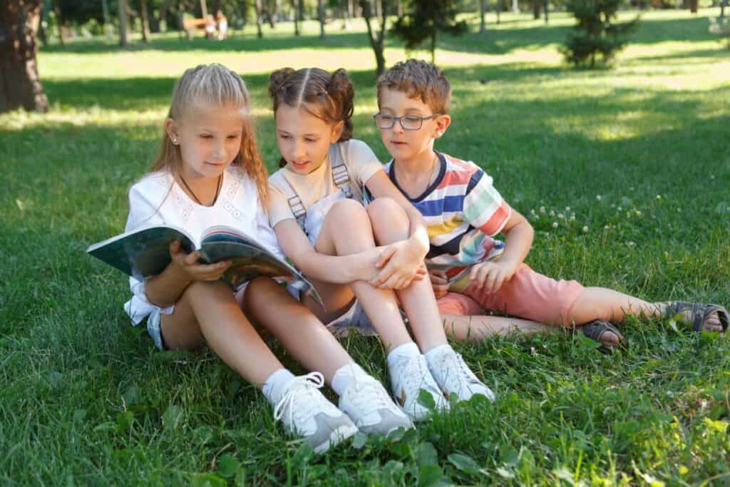 Three children sitting on the grass reading a book.