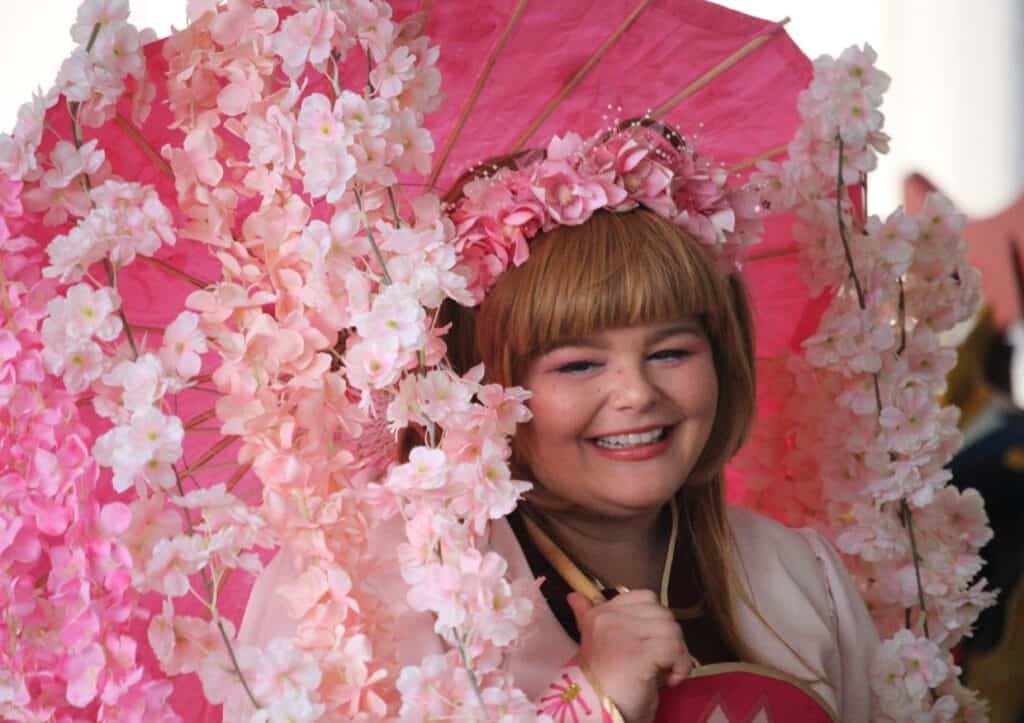 A woman smiling with pink flowers on her head, holding a pink umbrella at a place to see cherry blossoms.