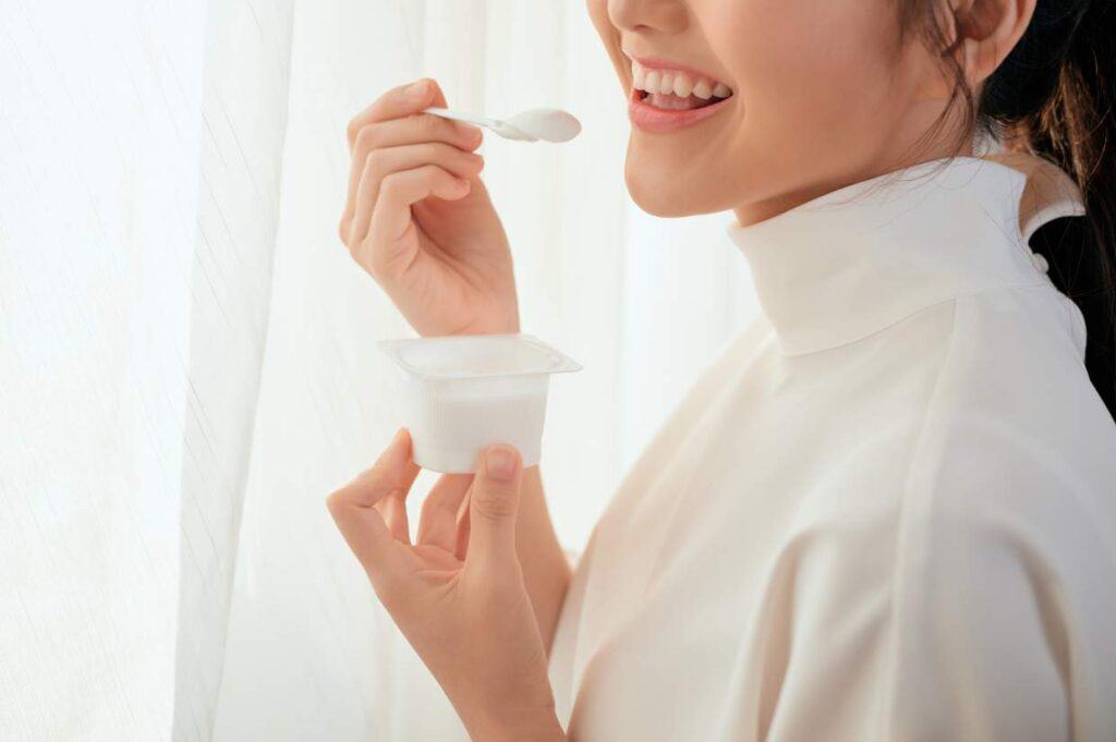 A woman is eating yogurt with a spoon in front of a window.