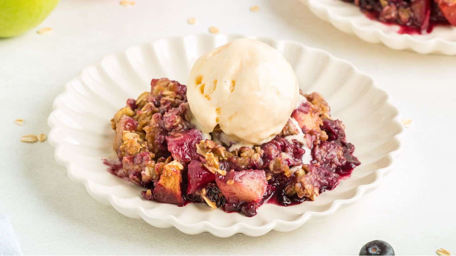 Apple and blueberry crumble on a white ribbed plate with a scoop of vanilla ice cream on top.