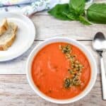 A bowl of red pepper and tomato soup next to a plate of bread and a spoon.