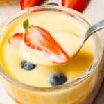 A spoon is being poured into a jar of lemon custard with strawberries and blueberries.