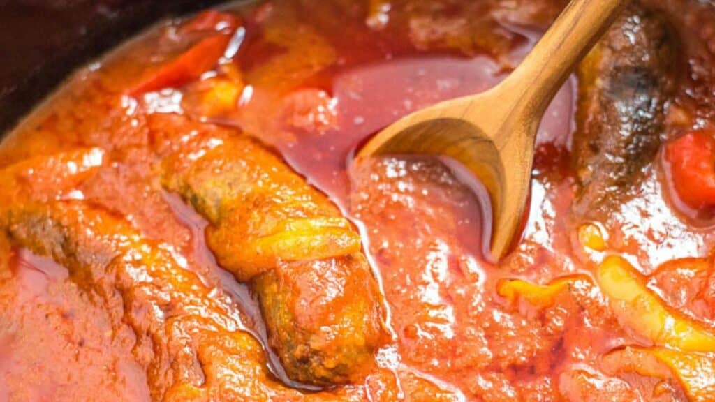 A pot of sausage, peppers, and marinara with a wooden spoon in it.