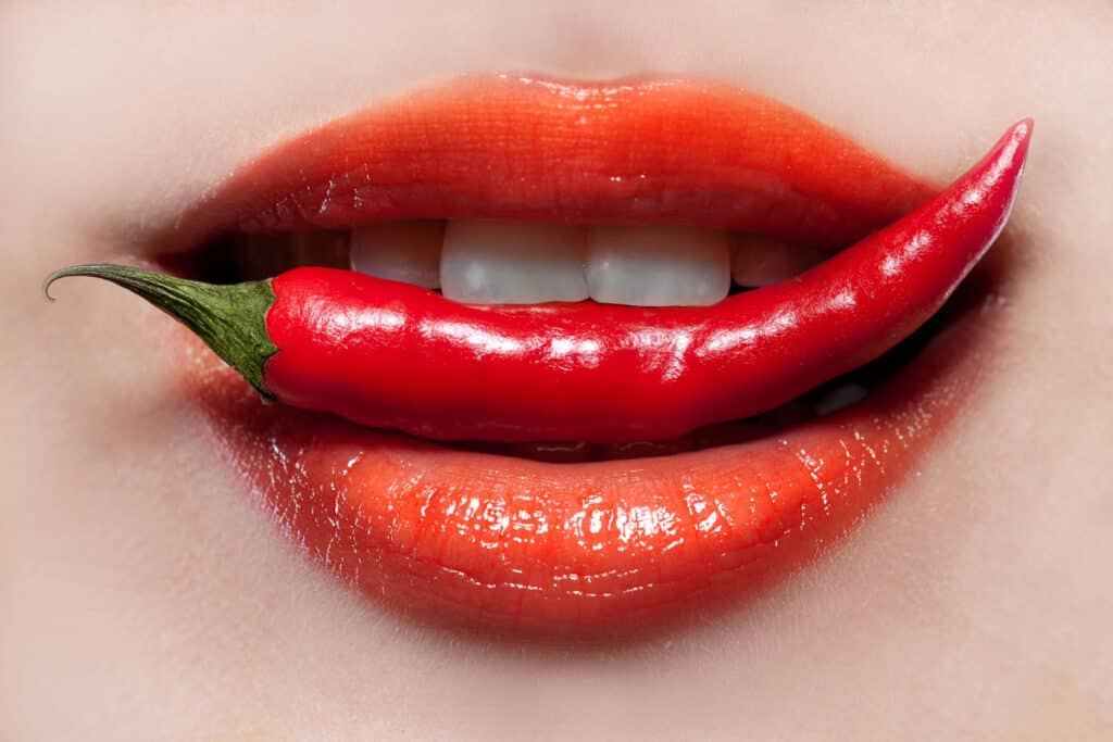 A close-up of a woman's mouth indulging in the spicy food trend with a red chili.