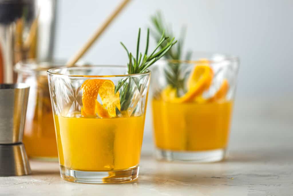 Two glasses of orange cocktail garnished with a slice of orange, a sprig of rosemary, and simple syrup substitutes.